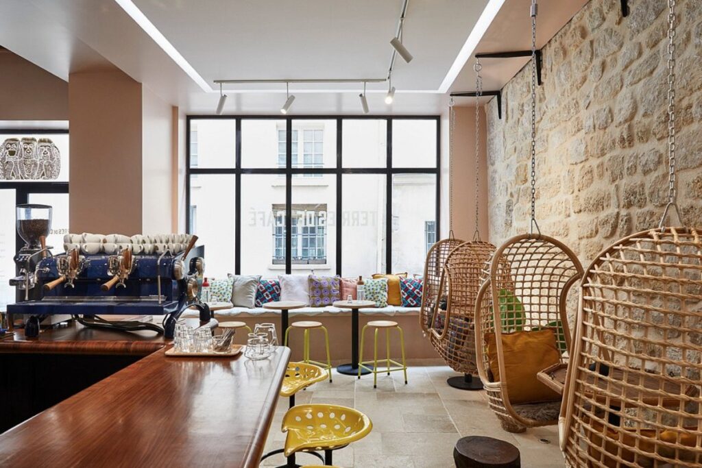 Terres de Café café interior with a cozy, inviting atmosphere. Features hanging rattan chairs, a stone wall, colorful plush pillows, and a sleek espresso machine.
