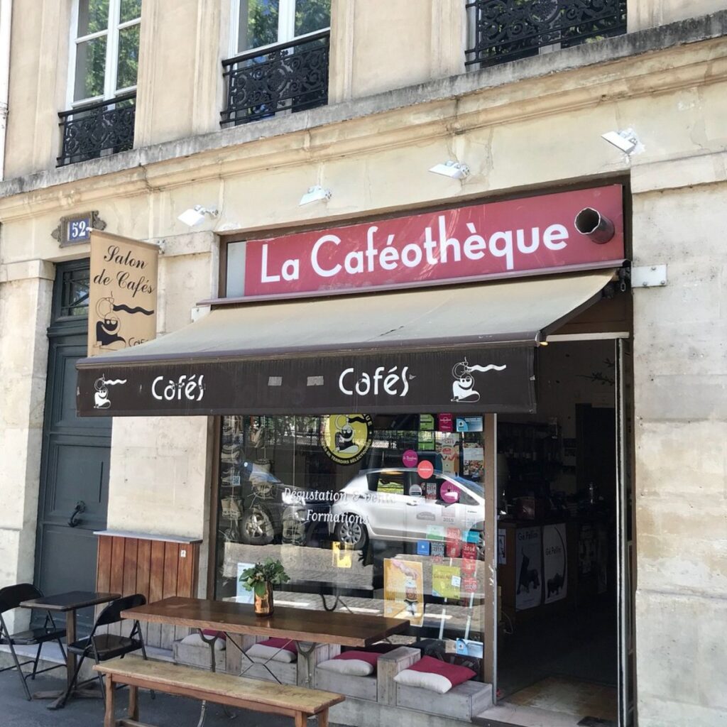 Street view of "La Caféothèque," a cozy cafe with a red sign, wooden bench, and dark awning. The inviting space features a glass storefront and outdoor seating.