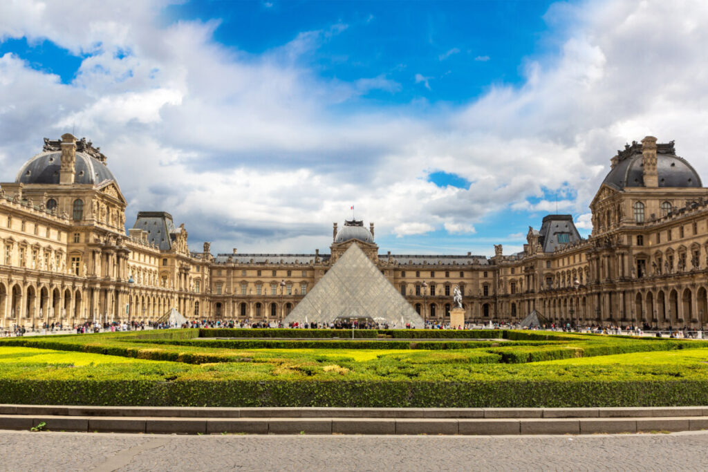 The Louvre Museum in Paris, with the iconic glass pyramid entrance, lush green lawns, and a vibrant blue sky with scattered clouds overhead.