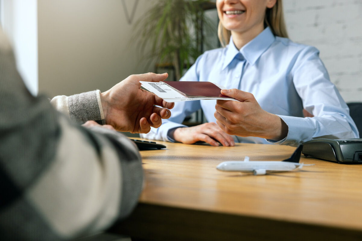 Travel agent in a light blue shirt smiles as she hands a passport and boarding pass to a customer at her desk.