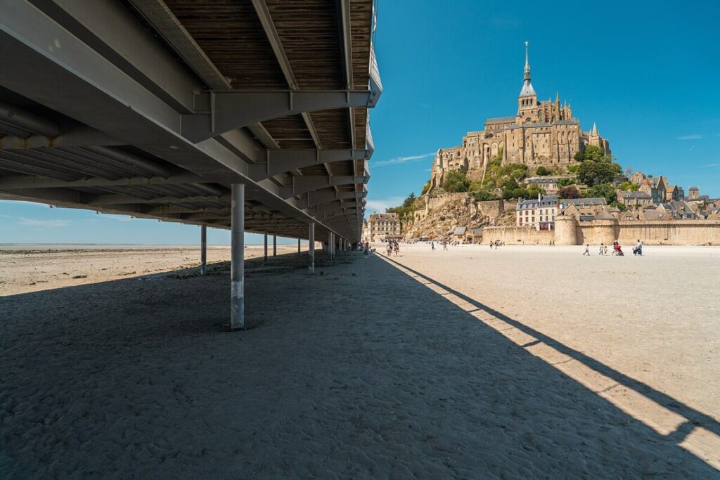 View of Mont Saint-Michel with bright blue sky, seen from beneath a bridge casting a long shadow on the sandy beach. People walk in the sun.