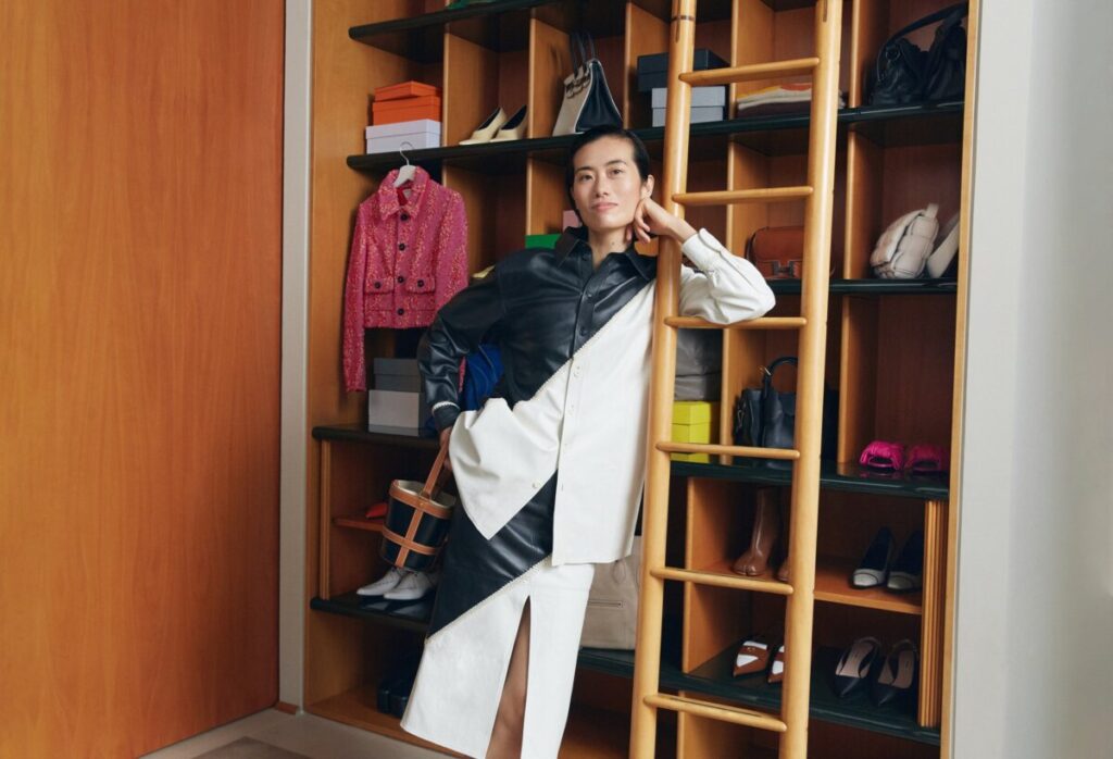 Woman wearing a white shirt and black jacket poses in front of a shoe display at Vestiaire Collective, Paris.