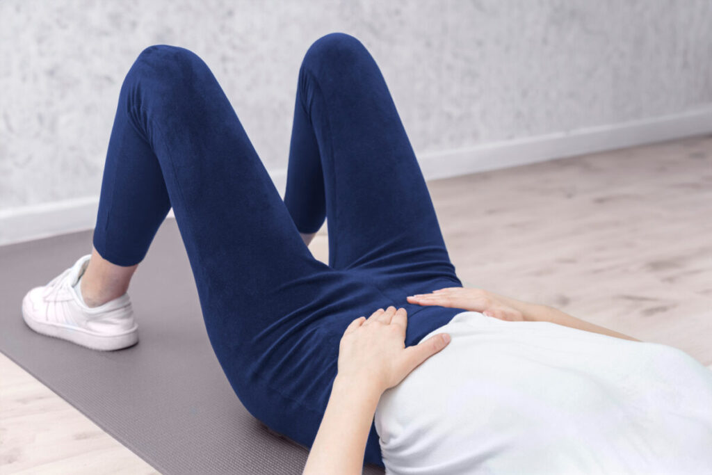 Person lying on a yoga mat, wearing navy leggings and white sneakers, with hands on abdomen. Relaxed indoor setting with a light-toned wooden floor.